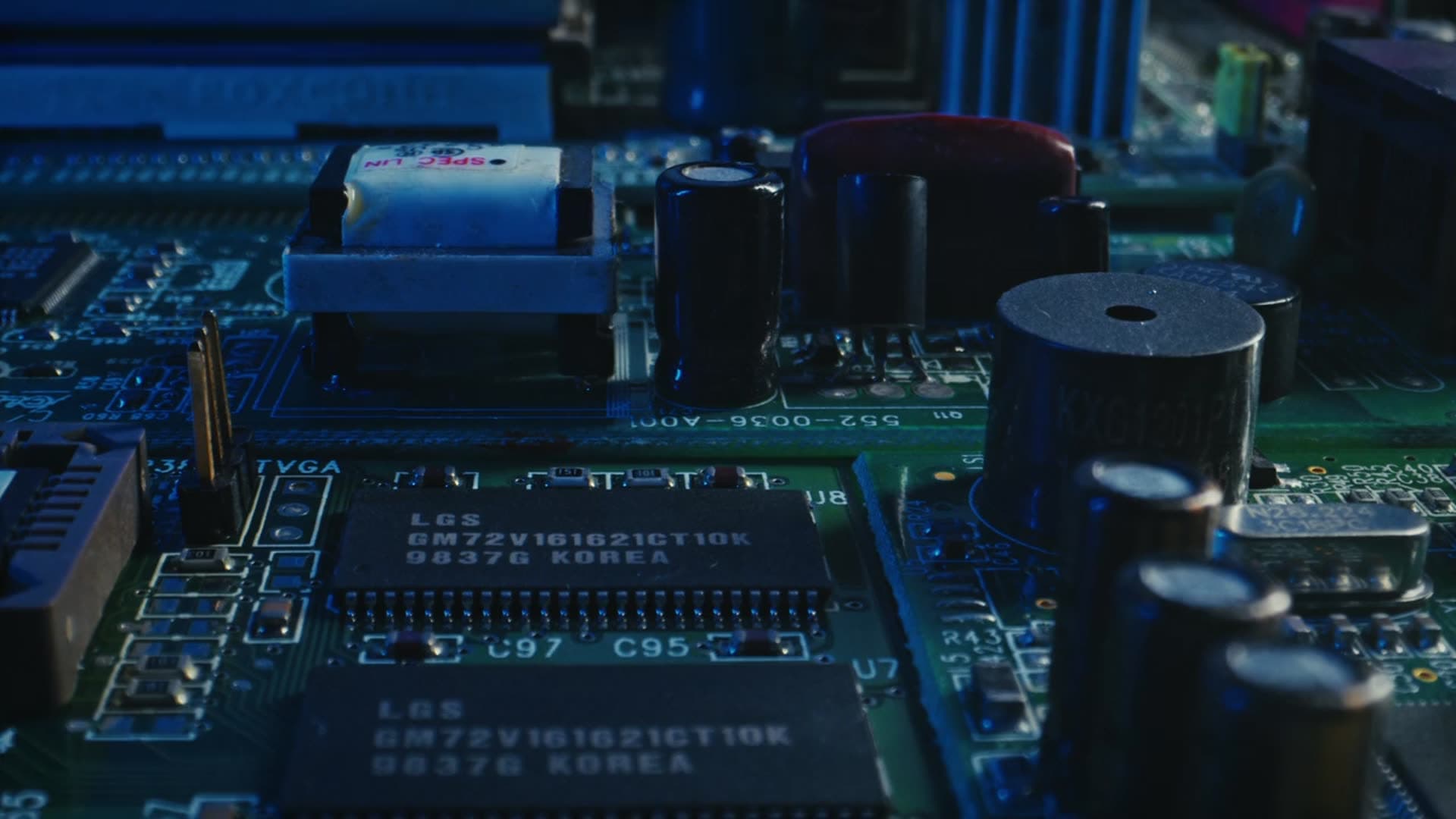 A dark green circuit board seen close up and at an angle, lit in blue, with memory chips, capacitors and a transformer in shallow focus.